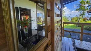 Una terraza de madera con barandilla de vidrio, una mesa de mimbre con sillas y vistas a un jardín exuberante y palmeras.