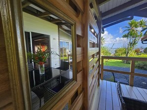 A wooden deck with a glass balustrade, a wicker table with chairs, and a view of a lush garden and palm trees. - Villa Fareatai (Raiatea)