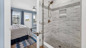 A bedroom with a bed, a shower with a glass door, and a view of buildings outside. - St. Somewhere in Seacliffs (Port St. Joe)