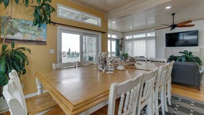 Un coin repas avec une table en bois, des chaises blanches, un ventilateur de plafond et un téléviseur fixé au mur.