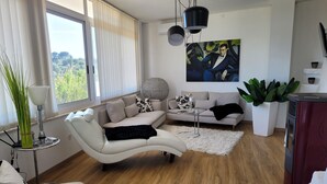 A modern living room with a white sofa, a black throw blanket, and a round coffee table. There is a large window with blinds, a painting on the wall, and a potted plant. - Trullo in panoramic position near Ostuni (Ostuni)