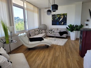 A modern living room with a white sofa, a black throw blanket, and a round coffee table. There is a large window with blinds, a painting on the wall, and a potted plant. - Trullo in panoramic position near Ostuni (Ostuni)