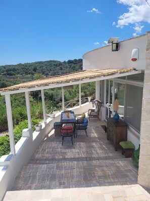 A covered patio with a table and chairs, a wooden cabinet, and a view of a hilly landscape. - Trullo in panoramic position near Ostuni (Ostuni)