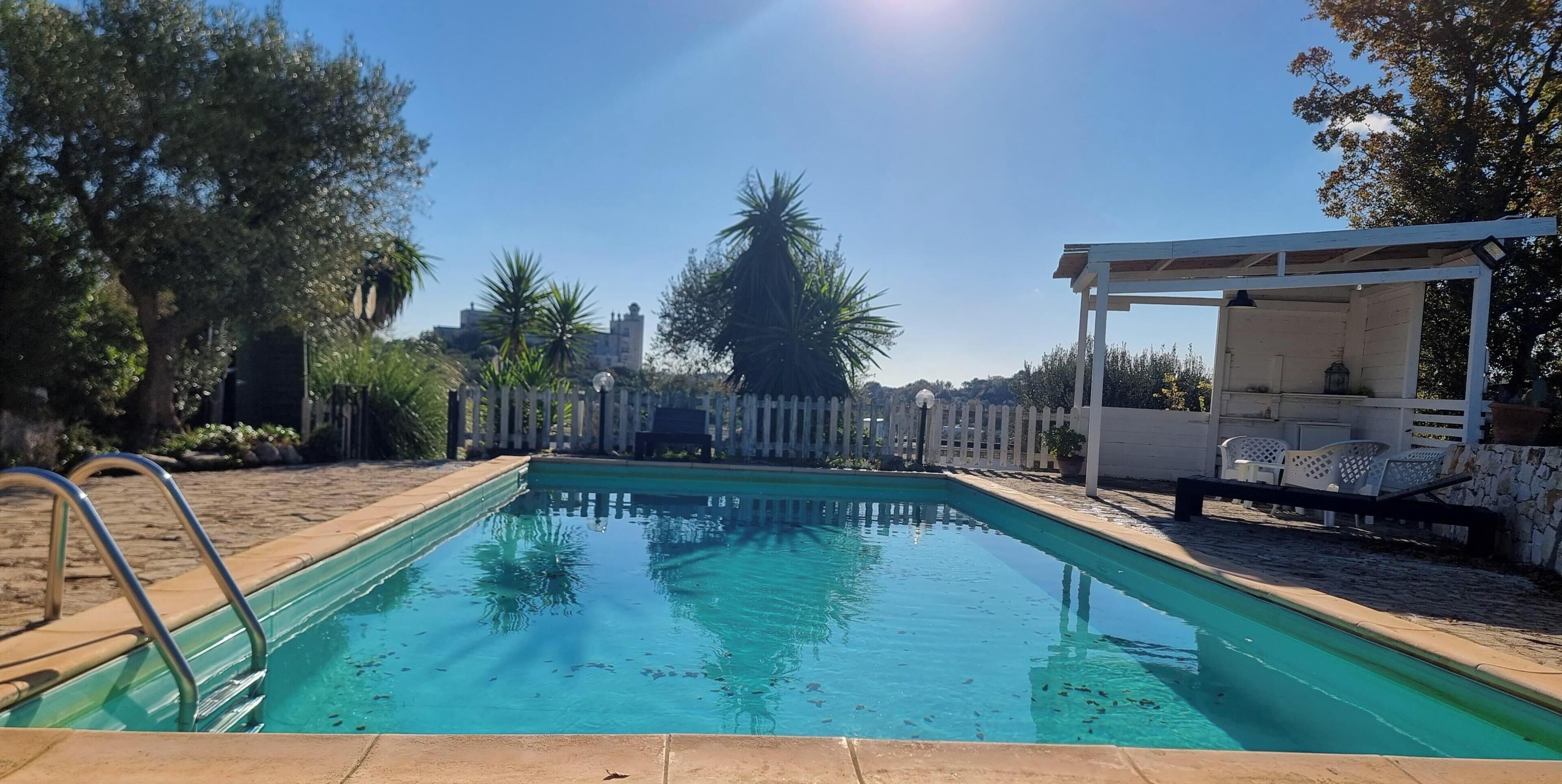A rectangular swimming pool with a metal ladder on one side, surrounded by a white fence and various trees.
