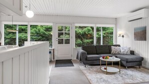 Ett modernt vardagsrum med en grå soffa, ett runt soffbord och utsikt över grönskan genom stora fönster.