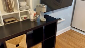 A kitchen area with a dark wooden table, white shelves with dishes, and a television mounted on the wall. - Modern, cozy unit in quiet neighborhood close to EVERYTHING. (Coral Gables)