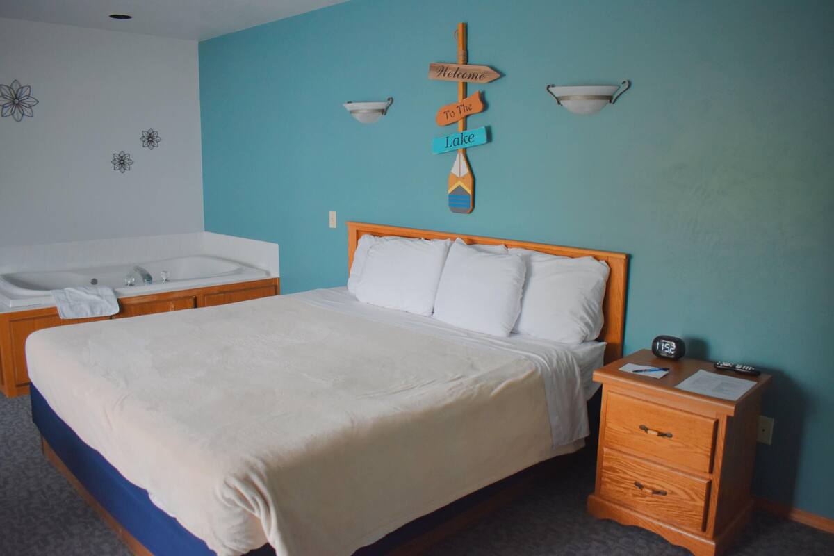 Un dormitorio con cama, mesita de noche, cabecera de madera, un letrero en la pared, bañera y una toalla.