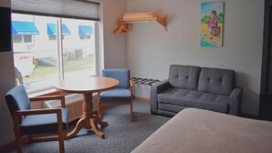 A hotel room with a bed, a grey sofa, a round table with two chairs, a window with blinds, a wall-mounted shelf, and a painting. - Innlet Motel (Sturgeon Bay)
