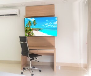 Ein moderner Schreibtisch mit Fernseher, der eine Strandansicht zeigt, ein schwarzer Bürostuhl und eine Holztischplatte.