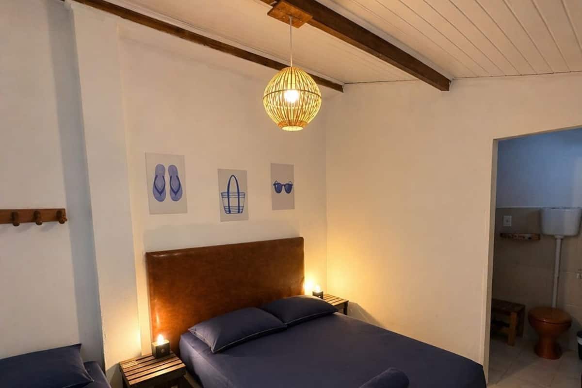 Un dormitorio con una cama grande, una mesita de noche, una lámpara colgante y un baño pequeño a la vista a través de una puerta abierta.