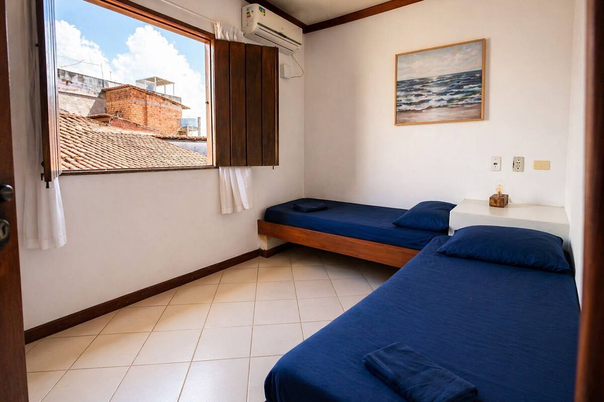 Una habitación con dos camas, una ventana que da a un edificio de ladrillo y un cuadro del mar en la pared.