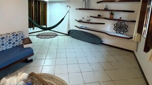 A living room with a sofa, a hammock, shelves with decorative items, and a patterned rug. - Nossa Casa Itacaré (Itacaré)