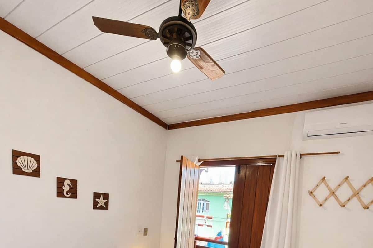 Un dormitorio con una cama, un ventilador de techo, una mesita de noche, una ventana con cortinas y vistas al exterior.