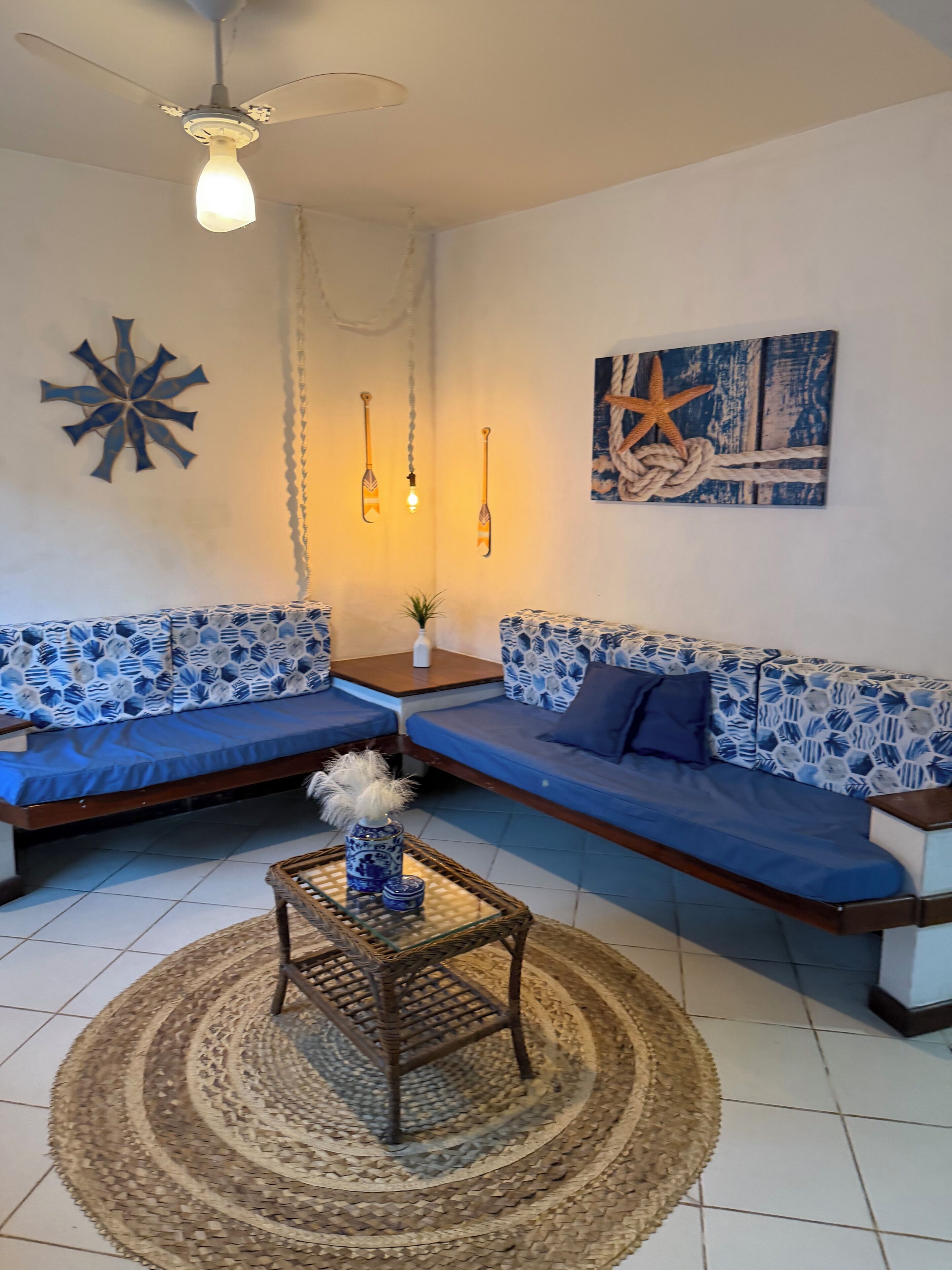 Una sala de estar con un sofá azul estampado, una mesa de centro redonda, un ventilador de techo y cuadros en la pared.