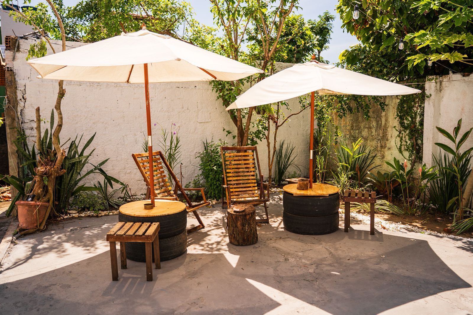 A patio area with two wooden chairs and two large umbrellas, surrounded by potted plants and a concrete floor.