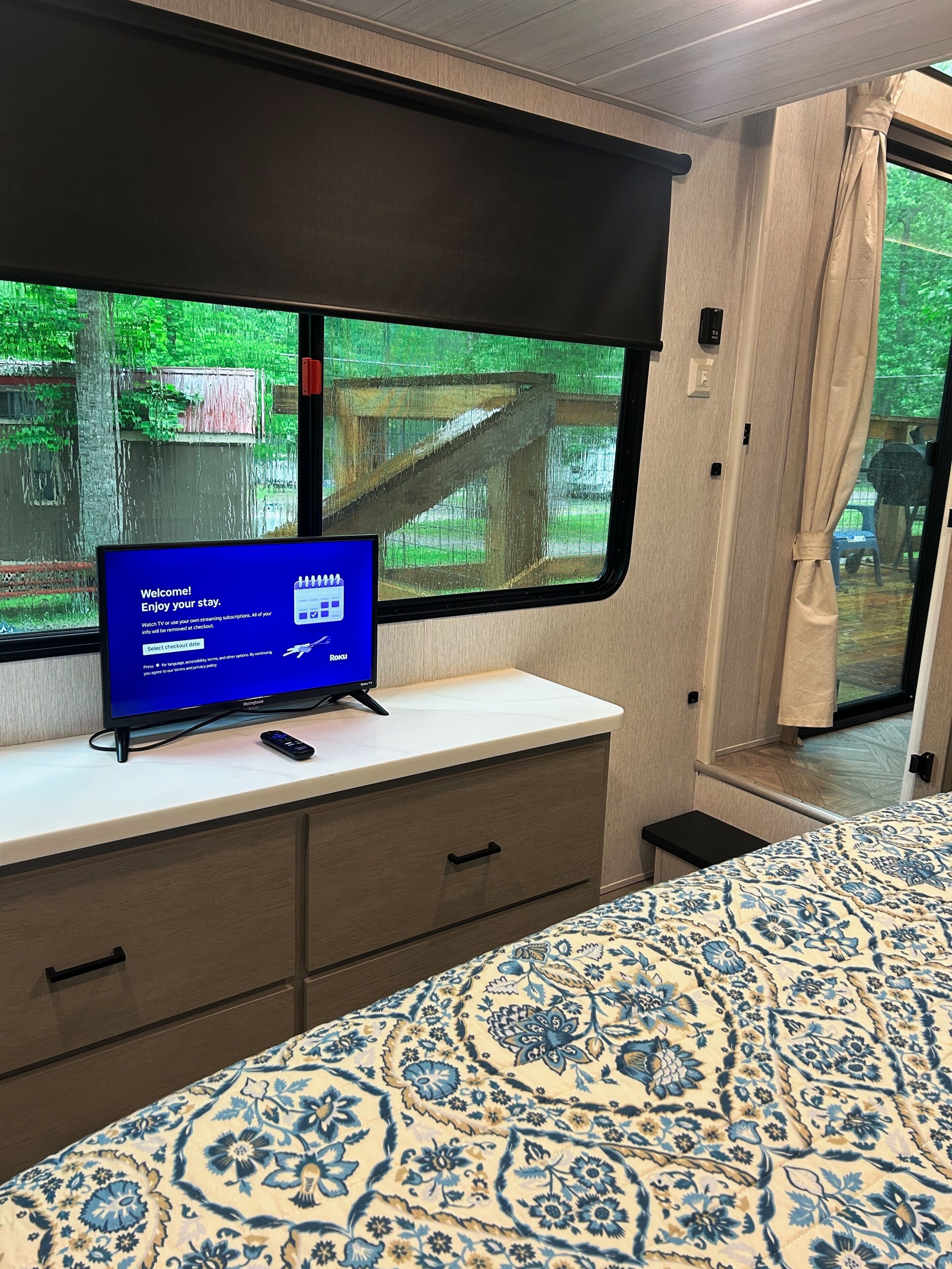 A room with a bed, a TV on a stand, a window with a view of greenery, and a curtain.