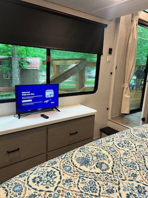 Ein Zimmer mit einem Bett, einem Fernseher auf einem Schrank, einem Fenster mit Blick ins Grüne und einem Vorhang.