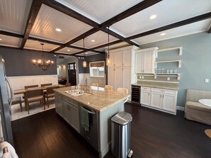 A modern kitchen with a central island, dark wood flooring, white cabinetry, and a dining area with a bench. - Refined Coastal Living Just Steps from Surf, Dining & Shopping (Huntington Beach)