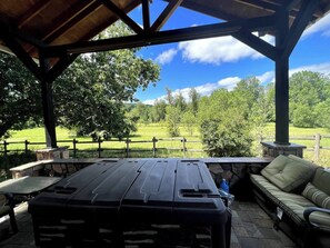 Um alpendre coberto ao ar livre com uma mesa de churrasco e assentos, com vista para um campo gramado e árvores.