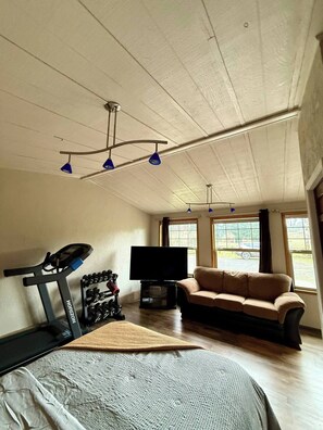 Quarto com ventoinha no teto, esteira, sofá, televisão e janelas amplas.