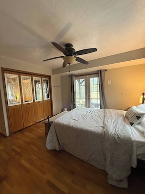 A bedroom with a bed, wooden wardrobe, ceiling fan, and large windows.