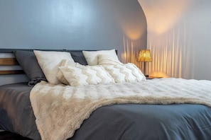 A modern bedroom with a curved headboard, a fluffy white throw blanket, and a bedside lamp. - T2 moderne et chaleureux en centre-ville (Lorient)