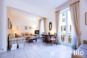 Ein Wohnzimmer mit einer Couch, einem Couchtisch und einem Esstisch mit Stühlen. Ein Fernseher steht auf einem Schrank, und ein Fenster ist mit Vorhängen drapiert.