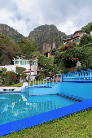 A swimming pool with a blue border, surrounded by lush greenery and a mountainous backdrop. - Hotel Bungalows Andrea (Tepoztlan)