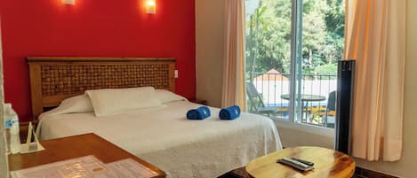 A hotel room with a bed, a wooden headboard, a window with curtains, a small table, and a chair.
