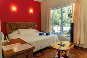 A hotel room with a bed, a wooden headboard, a window with curtains, a small table, and a chair. - Hotel Bungalows Andrea (Tepoztlan)