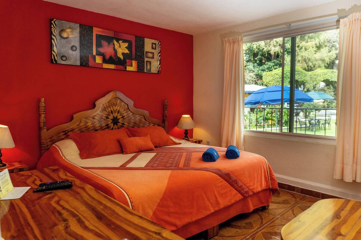 Habitación de hotel con cama, mesita de noche, lámpara, ventana con cortinas y vistas al exterior.