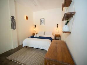 A bedroom with a bed, bedside lamps, a bedside table, a wall-mounted shelf, and a woven rug. - La Veranda & Restaurant  (Minca)
