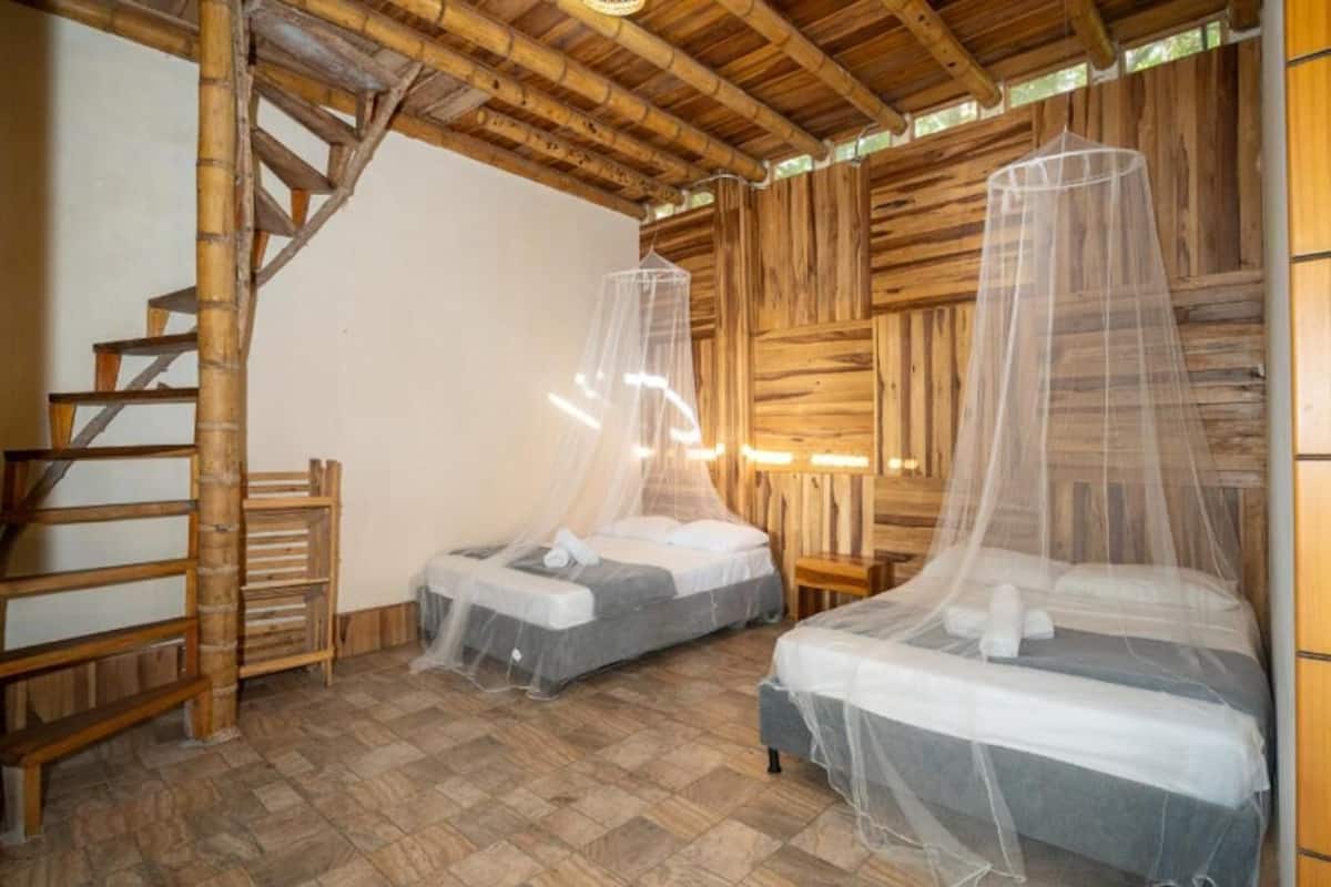 Habitació amb dos llits, cadascun amb mosquiteres, sòl de fusta, escala de fusta i paret de fusta rústica.