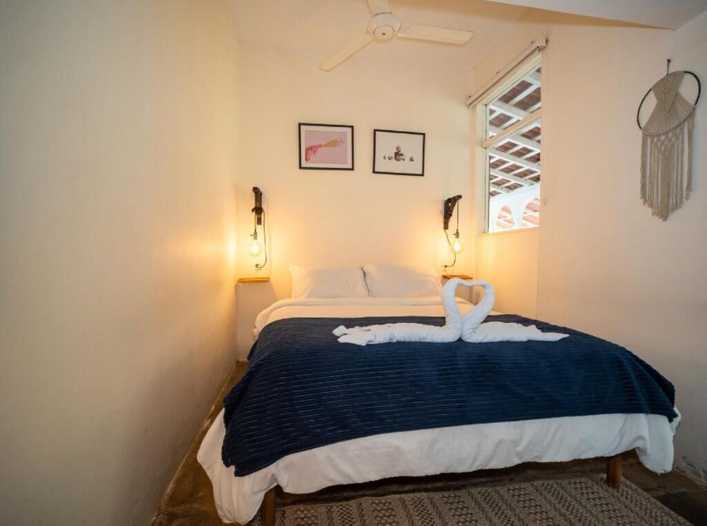 Un dormitorio con cama, dos lámparas de pared, ventilador de techo, un tapiz de macramé y dos cuadros enmarcados.