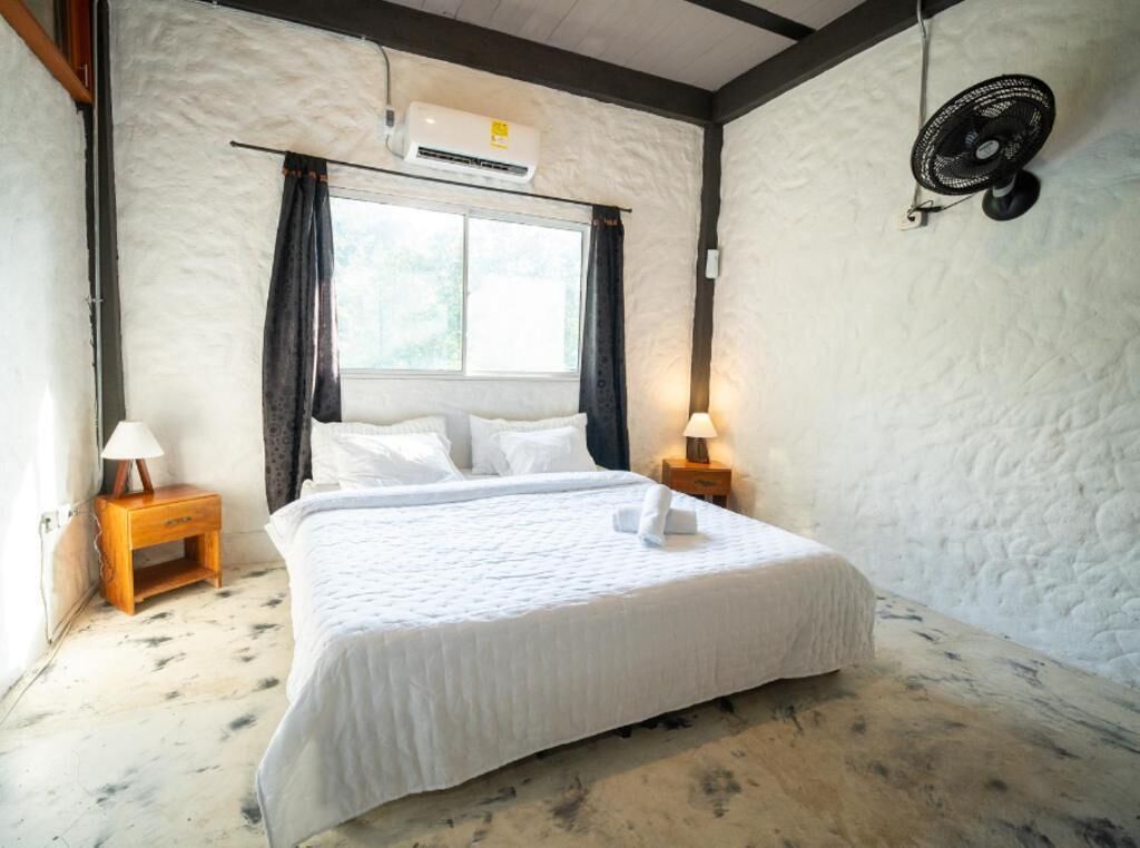 Un dormitorio con una cama, dos mesitas de noche, una ventana, un equipo de aire acondicionado y un ventilador de pared.