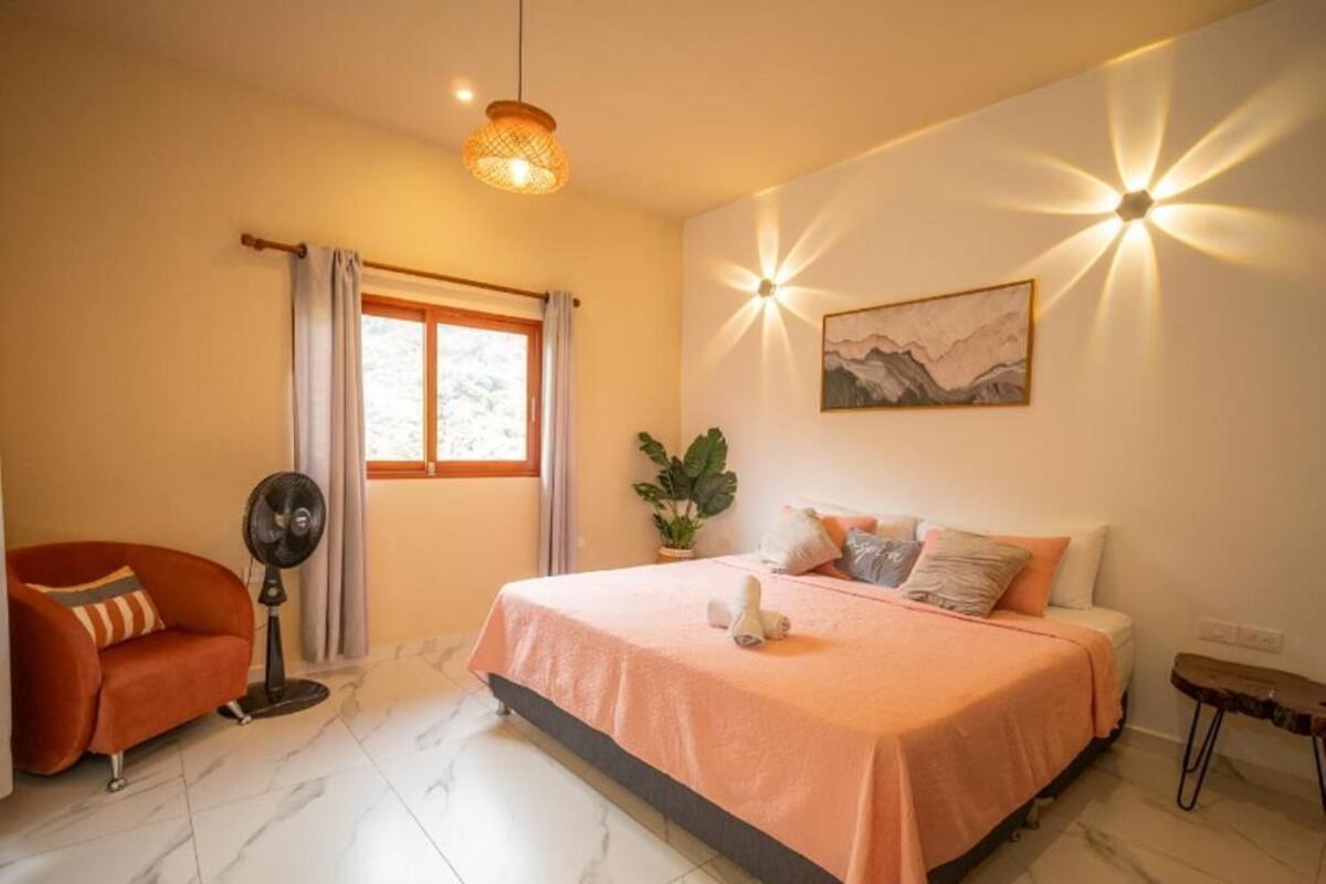 Un dormitori amb un llit, un ventilador, una cadira, una finestra i una làmpada penjada a la paret.