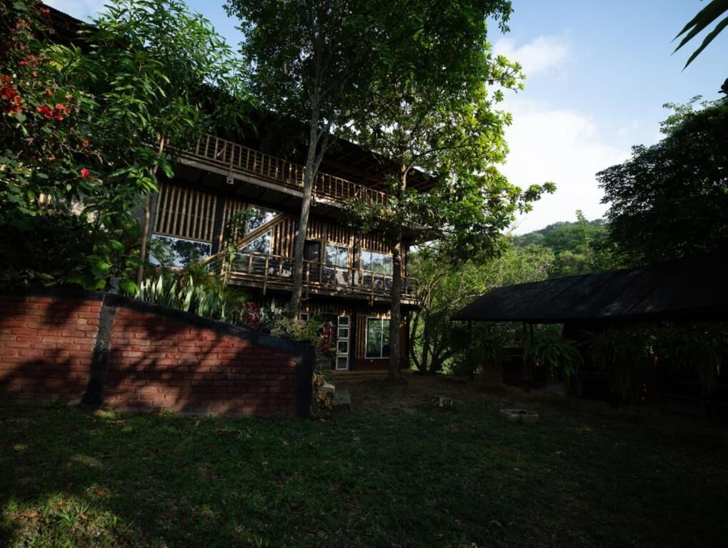 A two-story wooden structure with a balcony, surrounded by lush greenery and a brick wall.