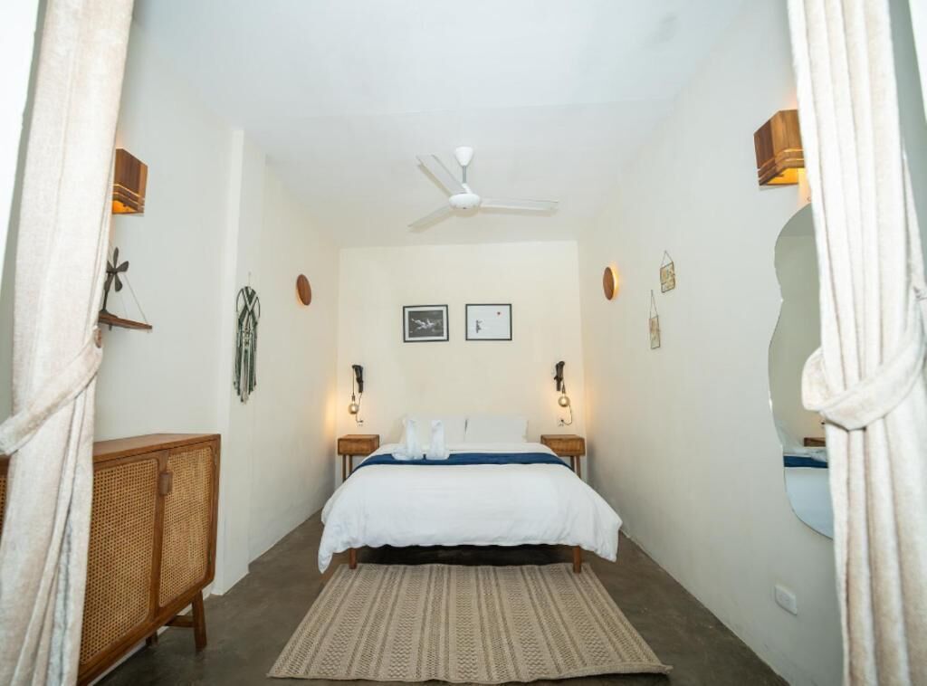 A bedroom with a bed, bedside lamps, a ceiling fan, a rattan side table, and a mirror.