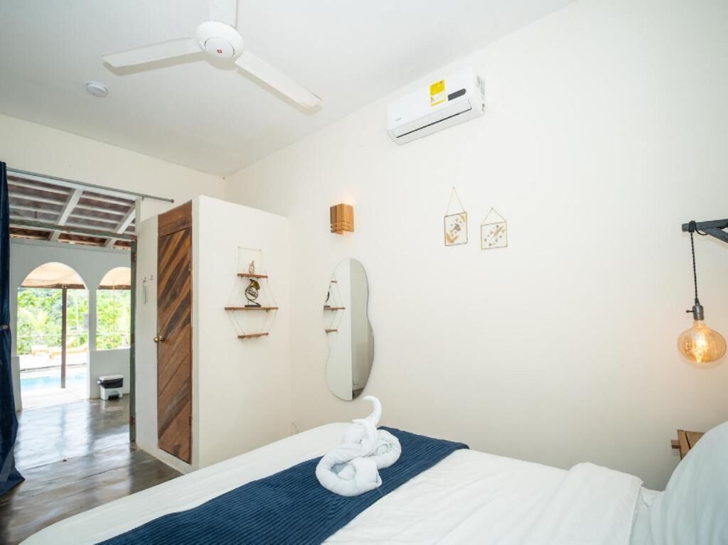 A hotel room with a bed, a ceiling fan, an air conditioning unit, a mirror, wall-mounted shelves, and a hanging light fixture.