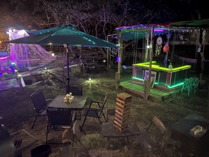 Un espace extérieur aménagé en terrasse avec des tables et des chaises, une structure en bois illuminée de néons et un espace de repos abrité.