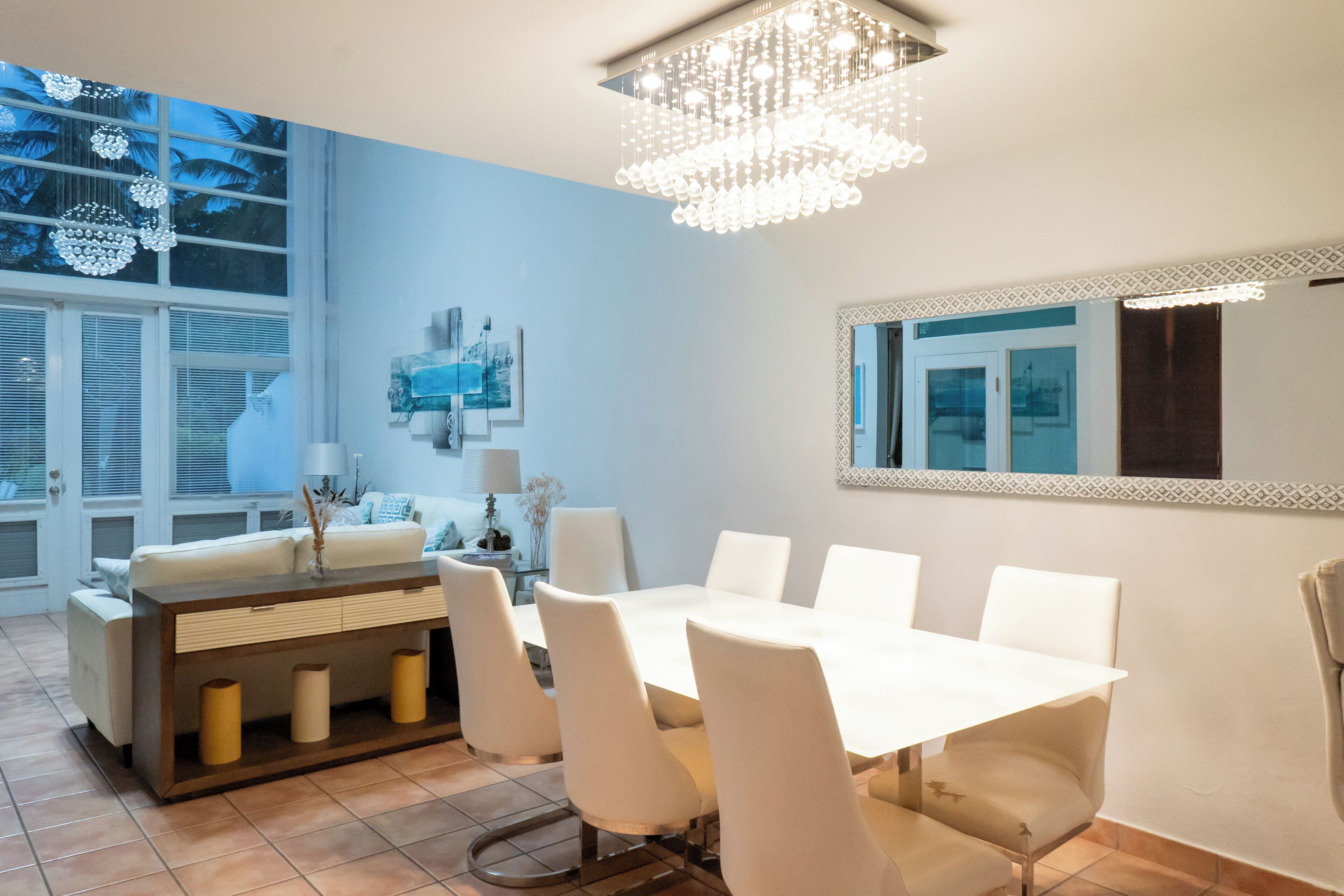 Un espace repas moderne avec une table et des chaises blanches, un lustre et une grande fenêtre équipée de stores.