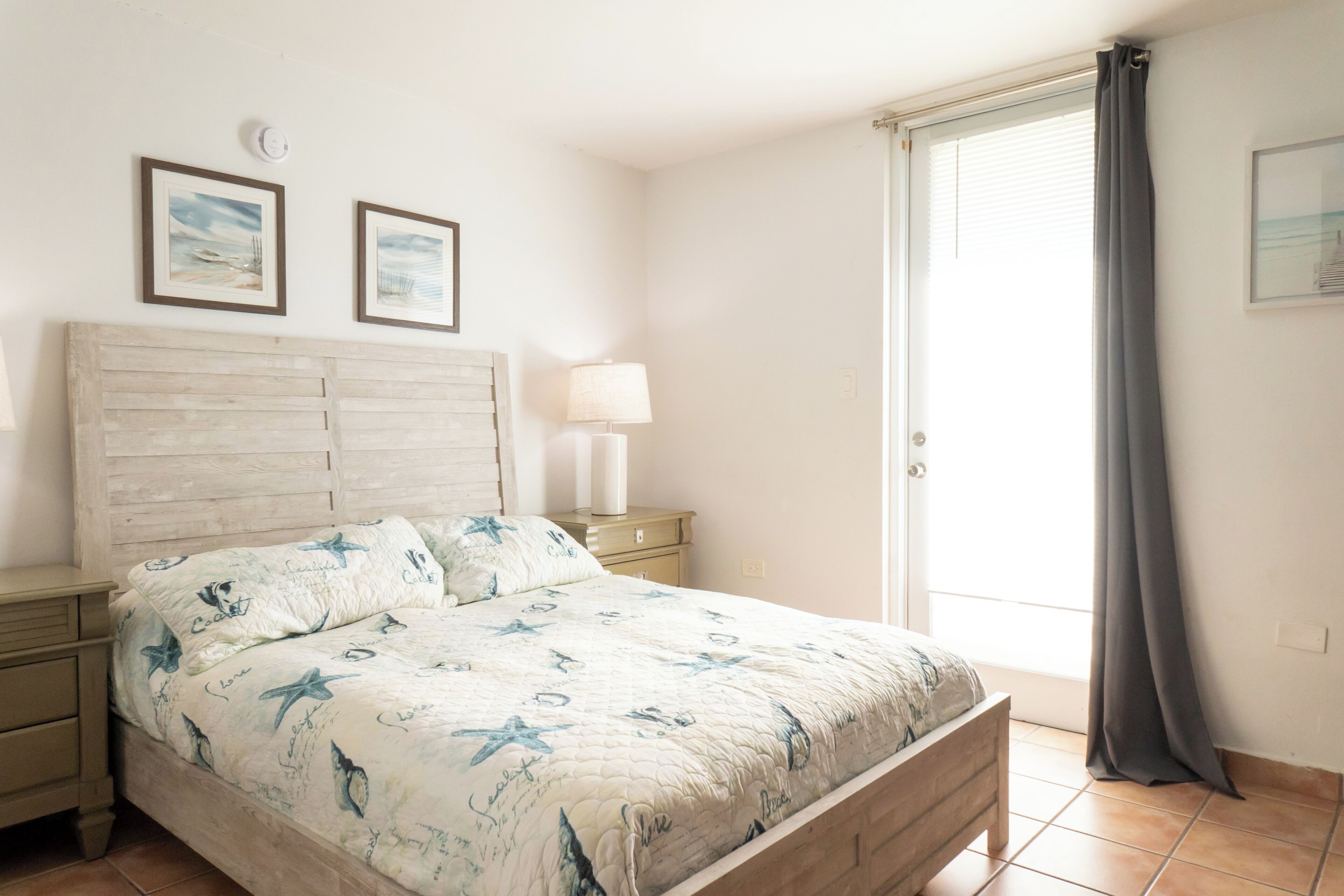 A bedroom with a bed, nightstands, a lamp, a window with curtains, and two framed pictures on the wall.
