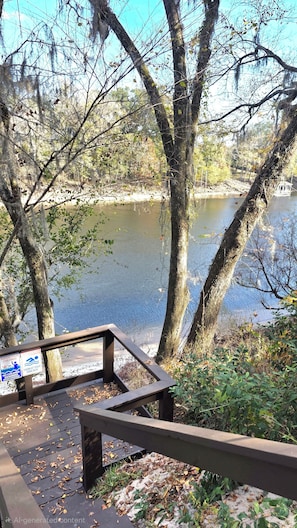 A wooden deck with a warning sign, overlooking a calm river with trees and a boat.