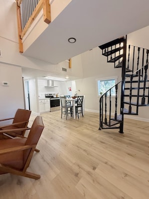 Amplia sala de estar con una escalera, una mesa de comedor con sillas y una cocina al fondo.