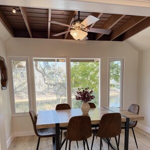 Comedor con mesa de madera, cuatro sillas, ventilador de techo y ventanas amplias con vista a árboles.