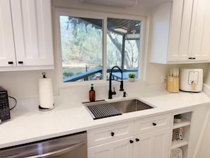 Una cocina con ventana, un lavamanos, un escurreplatos, un dispensador de jabón, un tostador y un bloque de cuchillos.