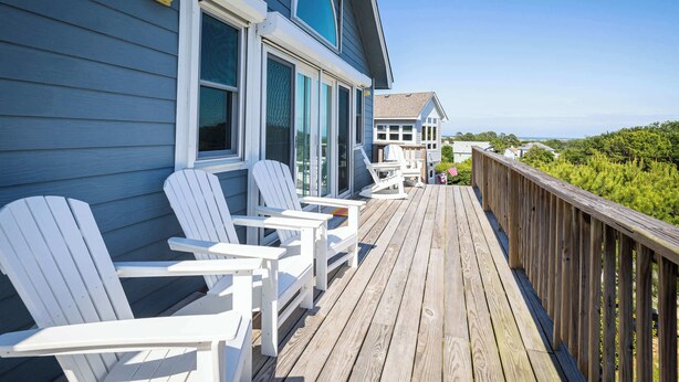 Una terraza de madera con sillas Adirondack blancas, vista al océano y una casa con revestimiento azul y ventanas grandes.