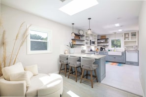 A modern kitchen with bar stools, a dining area with a sofa, and a large window. - Close to Beach & Duck Town w Pool & New Updates! (Duck)