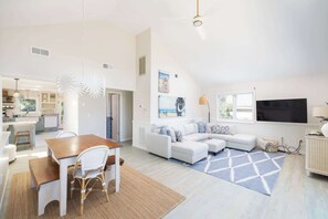 A modern living room with a white sofa, a dining table with chairs, a kitchen in the background, and a large window. - Close to Beach & Duck Town w Pool & New Updates! (Duck)