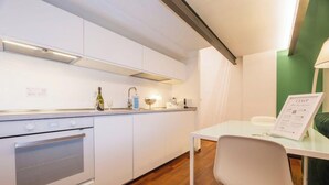 Cocina compacta con armarios blancos, horno empotrado, una mesa con una silla y una pared de acento verde.